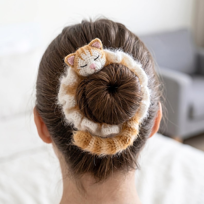 May include: A crocheted hair accessory in the shape of a sleeping cat. The cat is orange and white, with closed eyes and pink details. The hair accessory is wrapped around a bun, with a cream and orange striped pattern.