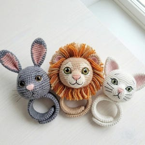 Può includere: Tre sonagli per bambini fatti a mano all'uncinetto a forma di animali: un coniglietto grigio con orecchie rosa, un leone con una criniera arancione e un gatto bianco. Ogni sonaglio ha una base ad anello. I volti sono ricamati.