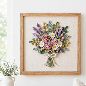 May include: A framed, handcrafted floral bouquet. The artwork features a variety of crocheted flowers in shades of pink, purple, yellow, and white, with green leaves. The bouquet is tied with a cream-colored bow and set against a neutral fabric background within a wooden frame.