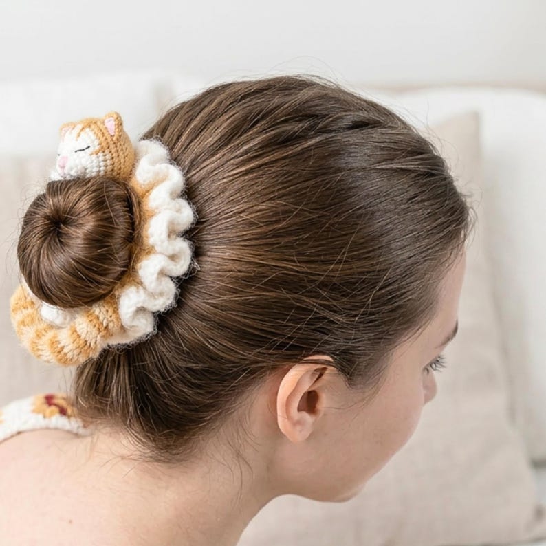 May include: A handmade hair scrunchie with a crocheted cat design. The scrunchie is cream and orange, with a ruffled edge. A small, sleeping cat is attached to the top of the bun, adding a whimsical touch.