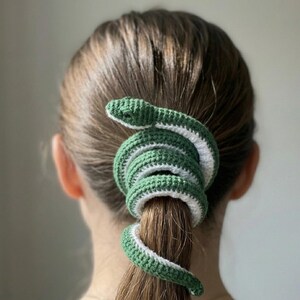 May include: A green and white crocheted snake hair accessory wrapped around a ponytail. The snake is green with a white stripe and a detailed head. The hair accessory adds a unique and whimsical touch.