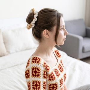 May include: A woman with brown hair wearing a cream, red, and yellow crocheted top and a matching scrunchie. The scrunchie and top feature a granny square design. The background includes a bed and a grey sofa.