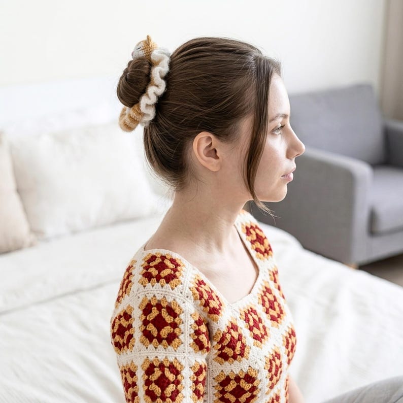 May include: A woman with brown hair wearing a cream, red, and yellow crocheted top and a matching scrunchie. The scrunchie and top feature a granny square design. The background includes a bed and a grey sofa.