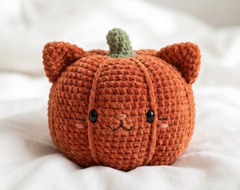 May include: A round, orange crochet pumpkin plush with two pointed cat-like ears on top and a small green stem. The plush features two black plastic eyes, a stitched nose, a mouth, and small pink embroidered cheeks on its textured surface.