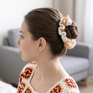 May include: A brown and white crocheted hair scrunchie with a cat design is worn in a bun. The cat has a light brown body and a white face. The scrunchie is made of soft yarn.