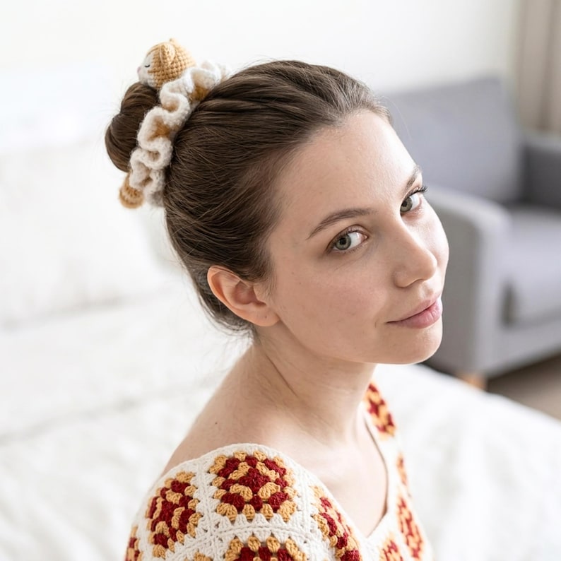 May include: A woman with brown hair in a bun, accessorized with a cream and tan crocheted scrunchie, topped with a small crocheted bear. She wears a cream and red crocheted top. The image is a close-up of the woman.