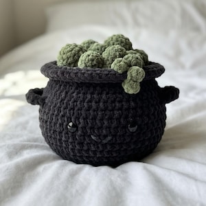 May include: A black crocheted cauldron with two side handles and a wide rim. The cauldron features two small black plastic eyes and a stitched mouth. Inside the cauldron are several round, textured, sage green crocheted spheres.