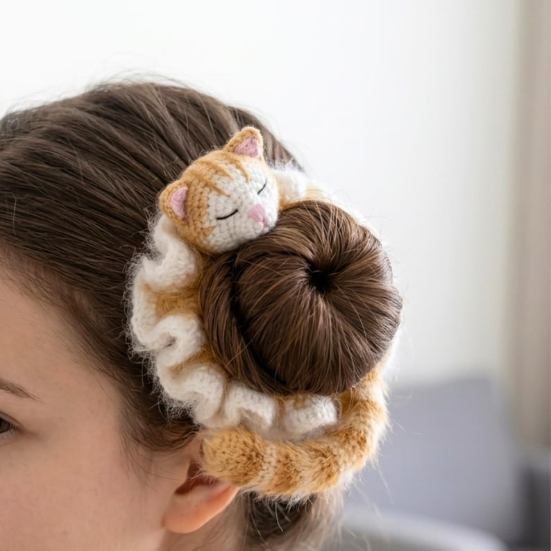 May include: A crocheted hair accessory featuring a sleeping ginger and white cat. The cat rests atop a bun of brown hair, surrounded by a ruffled, cream-coloured border. The cat's ears and nose are pink.