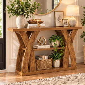May include: A wooden console table with an X-shaped frame and a lower shelf. The table holds a ribbed vase with greenery, a wooden bowl with spheres, books titled The Kinfolk At Home, a gold sculpture, two framed prints, and a table lamp.