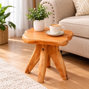 May include: A small, light-brown wooden side table with a natural, irregular edge top and three sturdy, thick legs. The table surface holds a white ceramic pot containing a green plant and a white ceramic coffee mug on a matching saucer.