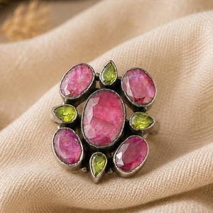 May include: A ring featuring a large central oval faceted pink stone surrounded by four smaller oval faceted pink stones and four small pear-shaped lime green stones, all set in a dark metal frame with a textured, oxidised finish.