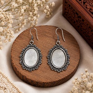 Può includere: Un paio di orecchini pendenti color argento con montature ovali e un bordo floreale decorativo. Ogni orecchino presenta un centro liscio e incavato e un semplice gancio per l'attacco, mettendo in risalto un design in metallo dettagliato.