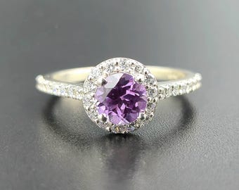 May include: A silver ring with a large, round, purple gemstone at the center, surrounded by a halo of smaller, clear stones. The band is also set with small, clear stones. The ring is displayed on a dark surface.