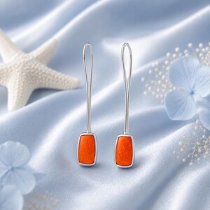 May include: A pair of long, slender silver drop earrings featuring a rectangular, vibrant orange stone at the base. The earrings have a minimalist, elongated wire design that extends upward from the stone, creating a sleek and balanced appearance.