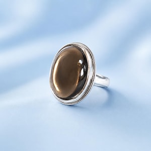 Op de afbeelding: Een zilveren ring met een grote, gladde, ovaalvormige rookbruine edelsteen gezet in een gepolijste metalen zetting. De steen heeft een reflecterend oppervlak met subtiele lichtreflecties en de ringband is van massief zilverkleurig metaal.