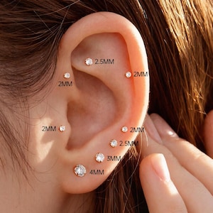 Può includere: Primo piano di un orecchio con diversi piccoli orecchini a bottone trasparenti. Le dimensioni sono indicate: 2 mm, 2,5 mm, 3 mm e 4 mm. L'orecchio è posto su uno sfondo sfocato, con una mano parzialmente visibile.