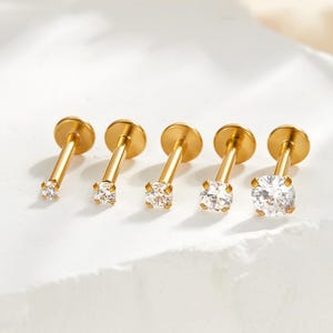 以下が含まれることがあります： クリア、丸、楕円形のキュービックジルコニアストーンが付いた、5つのゴールドカラーのリップピアススタッド。スタッドは白い表面に一列に並んでおり、さまざまなサイズとデザインを示しています。リップピアス用のジュエリーです。