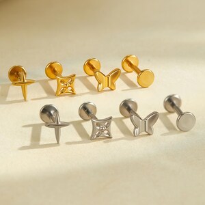 May include: A collection of gold and silver stud earrings. The earrings showcase diverse designs, such as a starburst, a geometric square, a butterfly, and a plain disc. The earrings are presented on a light beige surface.