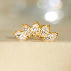 Può includere: Un orecchino color oro con un design a forma di ventaglio. L'orecchino presenta cinque gemme chiare, taglio marquise. Le gemme sono disposte in un motivo curvo, creando un aspetto delicato ed elegante. L'orecchino è posto su una superficie di colore neutro.