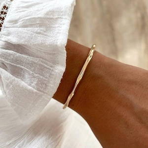 Può includere: Un bracciale rigido dorato con un design strutturato e organico. Il bracciale è indossato su un polso. Il design è leggermente irregolare, aggiungendo al suo fascino unico. Primo piano.