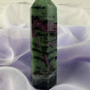 May include: A polished zoisite crystal tower, featuring green and red hues with a pointed top. The stone displays dark green and black bands with red streaks. The crystal stands upright against a soft, light purple fabric background.
