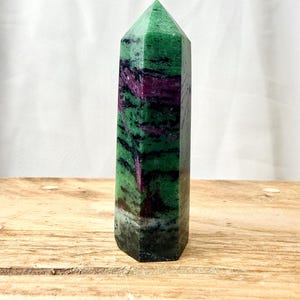 May include: A polished hexagonal crystal tower featuring a vibrant mix of deep green and magenta-purple mineral inclusions. The stone displays a pointed top and a smooth, multi-faceted surface with natural colour variations throughout the structure.
