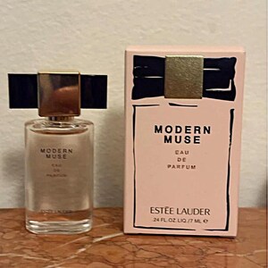 May include: A clear glass perfume bottle and its rectangular light pink box. Both feature the text Modern Muse Eau de Parfum and Estee Lauder. The bottle has a gold cap with a black bow design, and the box displays a matching black bow graphic.