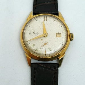 Może przedstawiać: Zegarek na rękę w stylu vintage w kolorze złotym z białą tarczą, złotymi wskazówkami i znacznikami. Zegarek posiada okienko datownika na godzinie 3 i czarny skórzany pasek. Na tarczy zegarka widnieje napis "Wastor 17 Jewels" i "anti-shock water-protected".