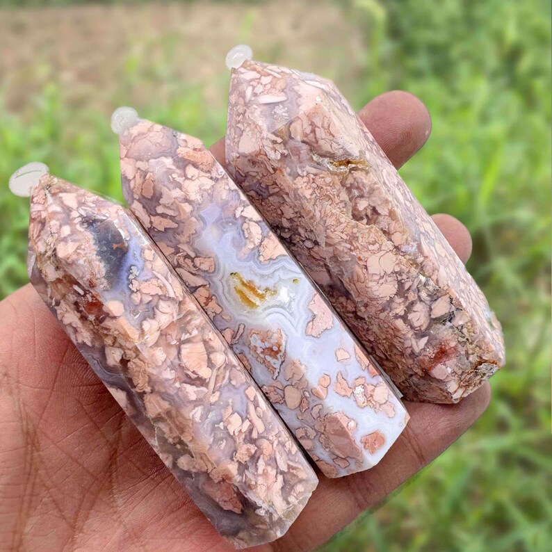 May include: Three polished gemstone wands with a mix of pink, brown, and white patterns. Each wand has a rounded top with a small, clear cap. The wands are held in a hand against a blurred green background.