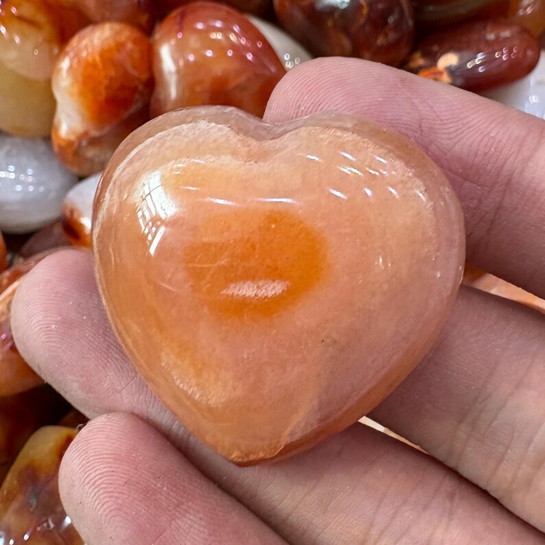 May include: A heart-shaped, polished gemstone in shades of orange and peach. The stone has a smooth, glossy surface and a lighter orange center. Other gemstones are visible in the background.