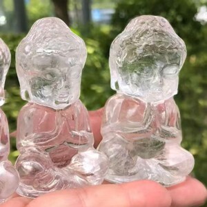 May include: Clear crystal Buddha figurines in a seated meditation pose. The figures are carved with detailed facial features and clothing, with hands clasped in prayer. The crystal is translucent, allowing light to pass through.