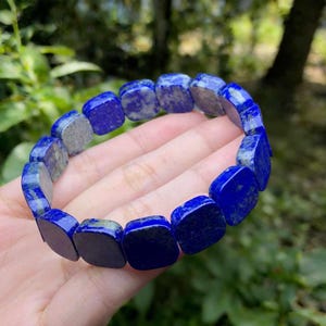 May include: A bracelet made of square-cut, deep blue lapis lazuli stones. The stones are strung together to form a flexible band. The bracelet is shown against a blurred green background.