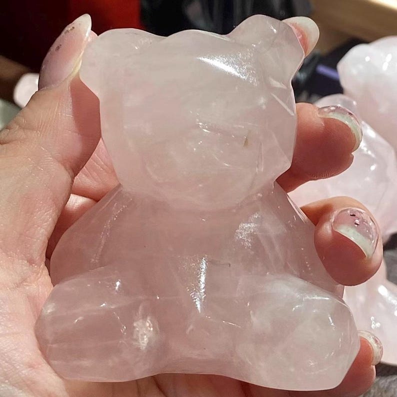 May include: A light pink rose quartz bear figurine. The bear is carved from a translucent stone, with visible natural variations in color. The bear is in a sitting position, with its arms and legs visible. The figurine is held in a hand.