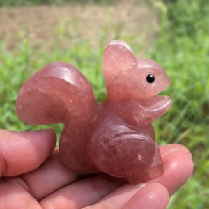 Puede incluir: Figurita de ardilla tallada en cuarzo rosa con un ojo negro. La ardilla está sentada con la boca abierta. La figura se sostiene en una mano sobre un fondo verde borroso.