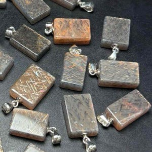 May include: A collection of rectangular gemstone pendants with silver-colored bails. The pendants have a mix of colors, including brown, gray, and orange, with a textured, crisscross pattern. The pendants are displayed on a dark surface.