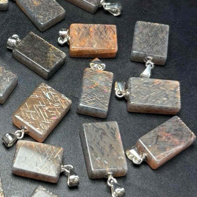 May include: A collection of rectangular gemstone pendants with silver-colored bails. The pendants have a mix of colors, including brown, gray, and orange, with a textured, crisscross pattern. The pendants are displayed on a dark surface.