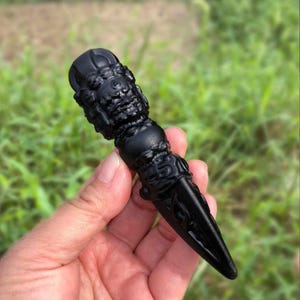 May include: A black, intricately carved ritual object with a detailed face and ornate patterns. The object has a conical shape with a pointed end. The background is blurred green grass.