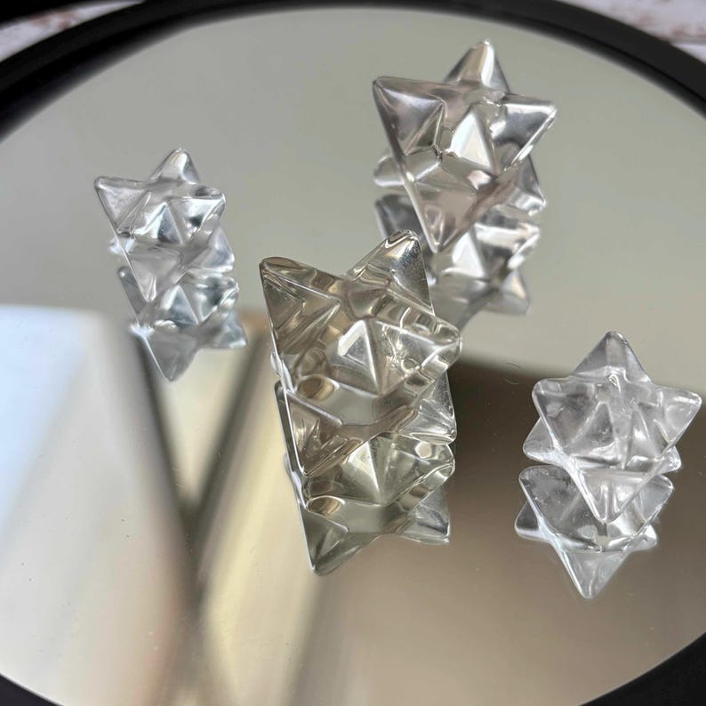 May include: Four silver-colored, geometric star-shaped objects are arranged on a reflective surface. The objects have a faceted design, catching the light and creating multiple reflections. The stars vary in size and are clustered together.