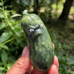 May include: A green, pickle-shaped sculpture with a carved, angry face and white, googly eyes. The sculpture is held in a hand against a blurred green background. The pickle has a textured surface and a small bump.