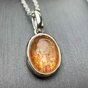 May include: A silver pendant necklace featuring an oval-shaped gemstone with a mix of orange, red, and gold hues. The stone is set in a silver bezel and hangs from a delicate silver chain. The pendant has a polished finish.