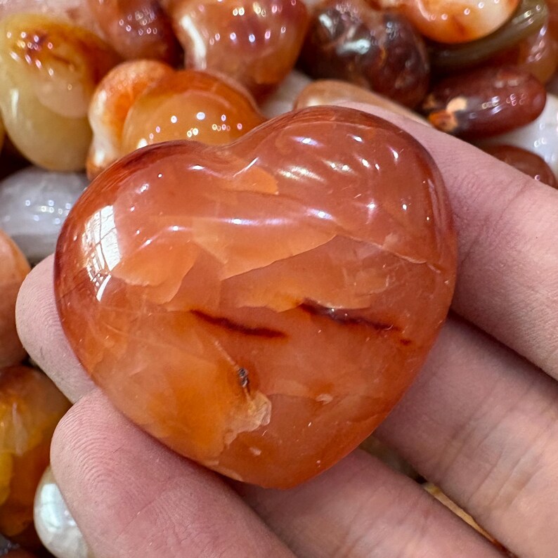 May include: A polished, heart-shaped carnelian stone, showcasing a vibrant orange-red hue with darker streaks. The gemstone is smooth and reflective, with a natural, organic appearance. Other carnelian stones are visible in the background.