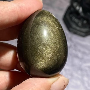 Op de afbeelding: Een gepolijste, eivormige gouden obsidiaansteen. De steen heeft een glad, reflecterend oppervlak met glinsterende gouden insluitsels. Het ei wordt tussen twee vingers gehouden, wat de grootte en vorm benadrukt. De achtergrond is wazig.