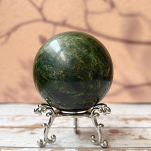 Peut inclure: Une sphère de pierre précieuse verte polie, avec un marbrage vert foncé et clair. Elle est présentée sur un présentoir décoratif en métal argenté avec des pieds ornés. L'arrière-plan est flou et de ton neutre.