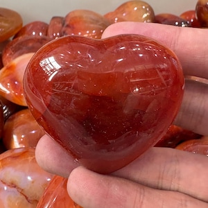 May include: A polished, heart-shaped carnelian stone. The stone is a deep reddish-orange color with a smooth, reflective surface. Other carnelian stones are visible in the background. This heart-shaped stone is a decorative item.