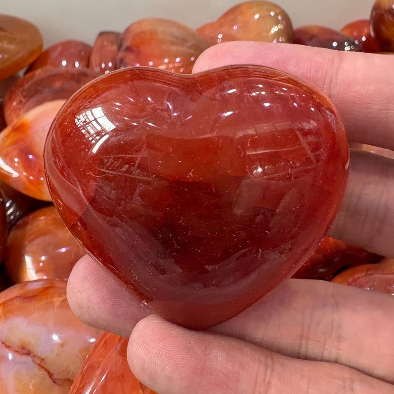May include: A polished, heart-shaped carnelian stone. The stone is a deep reddish-orange color with a smooth, reflective surface. Other carnelian stones are visible in the background. This heart-shaped stone is a decorative item.