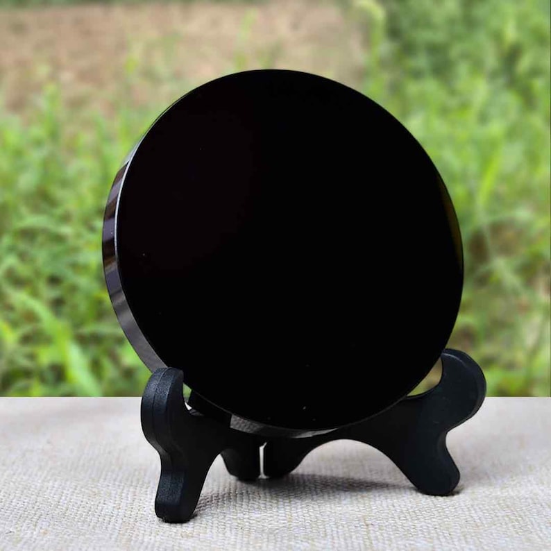 K&ouml;nnte beinhalten: Ein polierter, runder, schwarzer Obsidianstein, der auf einem kleinen, schwarzen Holzst&auml;nder pr&auml;sentiert wird. Der Stein hat eine reflektierende Oberfl&auml;che, und der St&auml;nder hat ein dreibeiniges Design. Der Hintergrund ist verschwommen und zeigt gr&uuml;nes Laub.