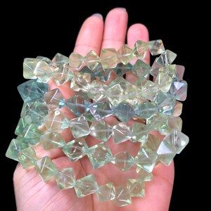 May include: A strand of faceted fluorite beads in shades of green and blue. The beads are square-shaped with a slightly translucent appearance, creating a sparkling effect. The beads are held in a hand against a black background.
