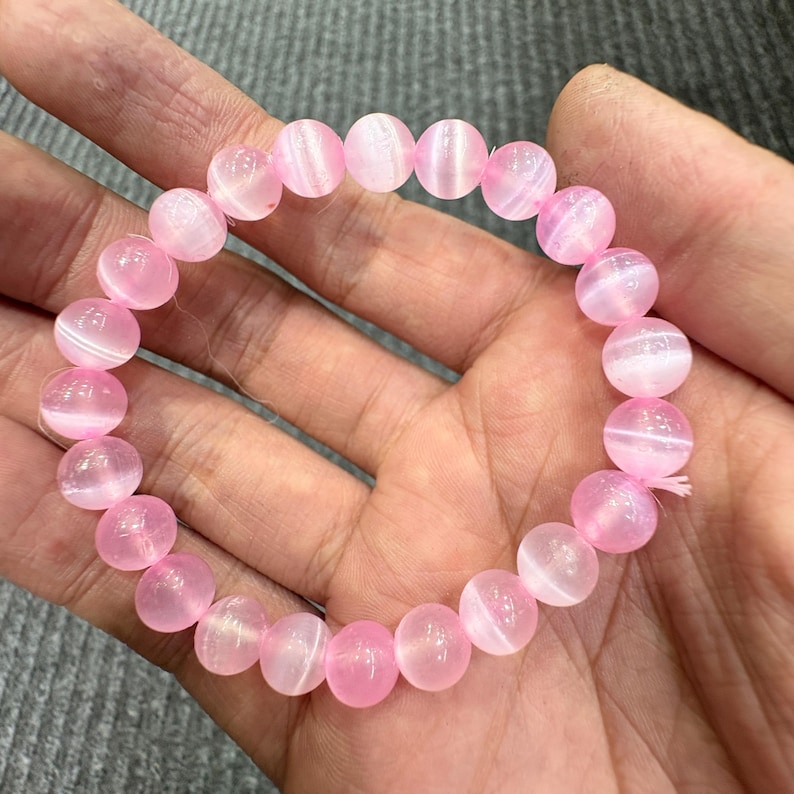 May include: A pink beaded bracelet with round, polished beads. The beads have a translucent quality with white striations. The bracelet is strung on a pink elastic cord. The bracelet is shown on a person's hand.