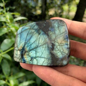 May include: A polished labradorite stone with a mix of blue, teal, and black colors. The stone has a unique pattern of lines and cracks, and is held in a hand. The stone is a natural gemstone.