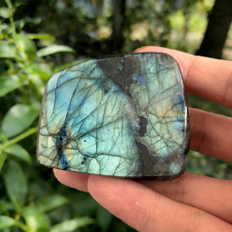 May include: A polished labradorite stone with a mix of blue, teal, and black colors. The stone has a unique pattern of lines and cracks, and is held in a hand. The stone is a natural gemstone.